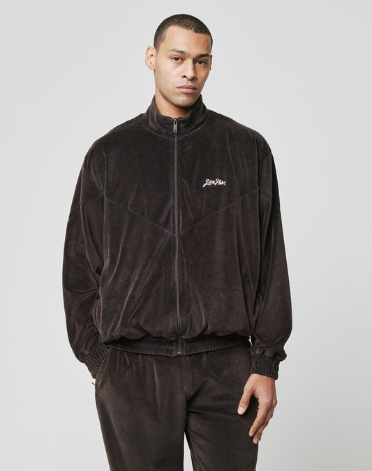 A man stands against a plain background wearing the LIVE FAST DIE YOUNG LFDY Velour Jacket in dark brown, featuring an oversize fit, zip-up front, subtle left chest logo, and matching pants.