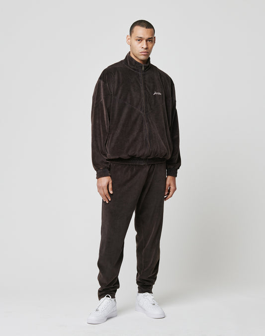 A man wears the LIVE FAST DIE YOUNG LFDY Velour Jacket and matching pants in an Oversize Fit, with a Stehkragen and subtle logo stitch, paired with white sneakers. He stands against a plain background, arms relaxed by his sides.