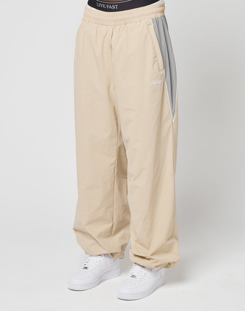 A person in LIVE FAST DIE YOUNG Basic Nylon Trackpants (wide fit, beige with light gray side panels, zip pockets, elastic drawcord) and a black LIVE FAST waistband stands against a plain white background, wearing white sneakers.