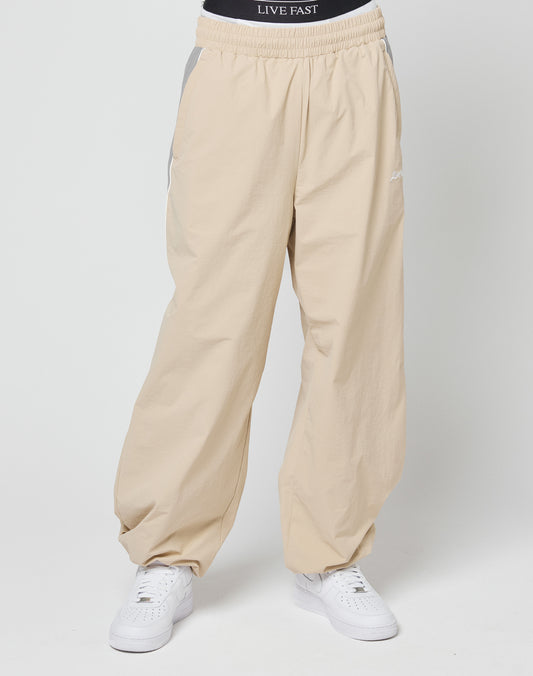 A person wears beige Basic Nylon Trackpants from LIVE FAST DIE YOUNG, featuring an elastic waistband and white sneakers. The black LIVE FAST DIE YOUNG underwear’s white logo is visible above the pants. Background is plain and light-colored.