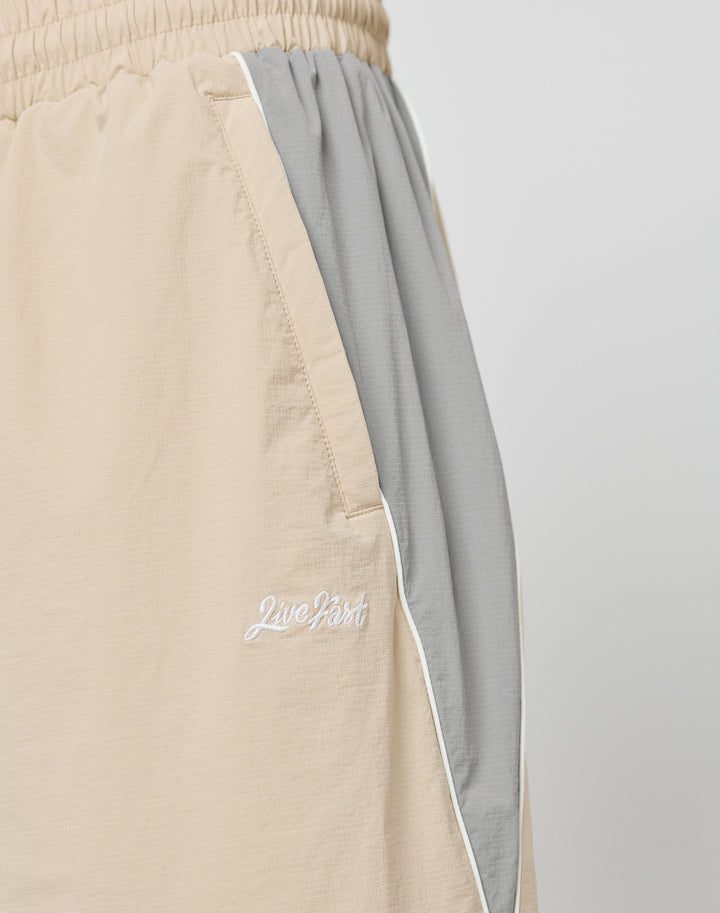 Close-up of beige and gray Basic Nylon Trackpants by LIVE FAST DIE YOUNG, featuring a wide fit, elastic waistband with drawcord, side pocket, white piping, and Live Fast embroidered in white on the left leg.