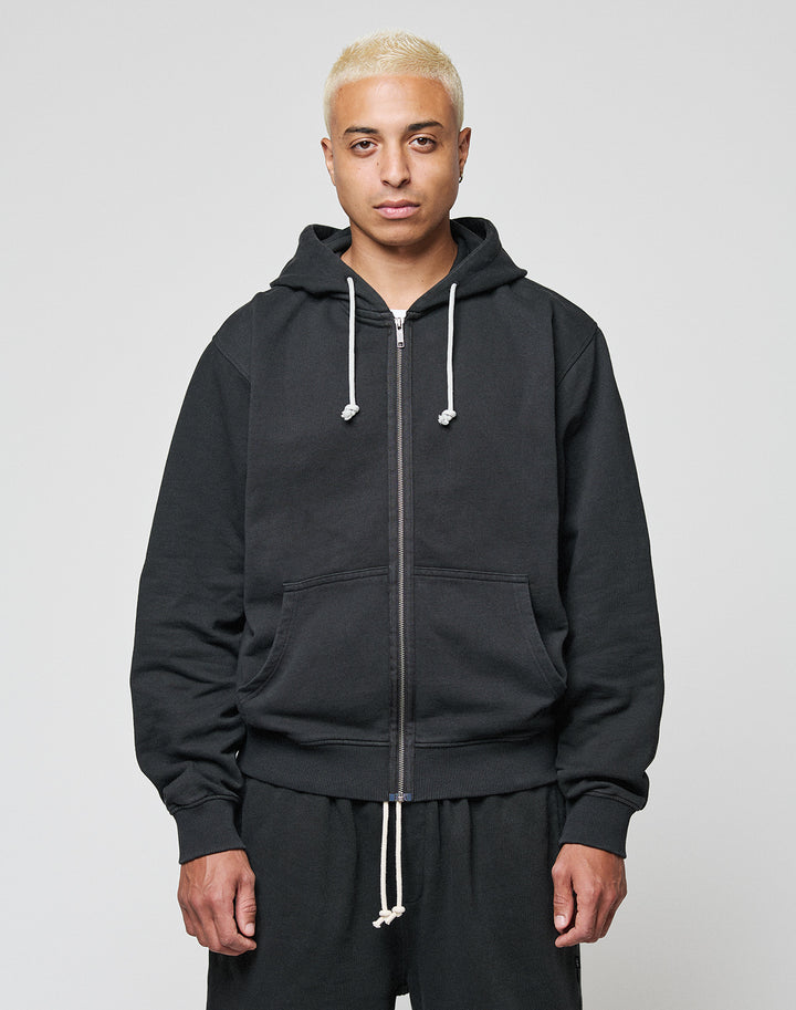 A person with short blond hair wears a black LIVE FAST DIE YOUNG LFDY Lightweight Hooded Zip and matching black pants, standing facing the camera against a plain light background.