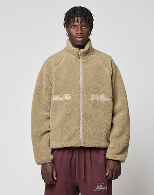 A man wears the tan LFDY Teddy Jacket by LIVE FAST DIE YOUNG, with Oversize Fit and zipper, featuring "Live Fast and Die Young" embroidery on the front. He pairs it with maroon sweatpants against a plain light gray background.