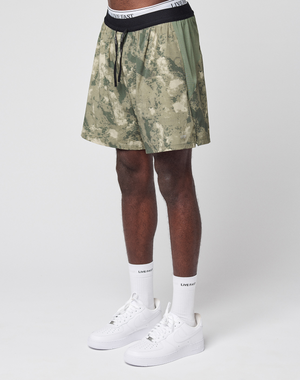 A person wearing green camo LFDY Performance Running Shorts by LIVE FAST DIE YOUNG, featuring a white LIVE FAST waistband, white socks, and white Nike sneakers stands against a plain white background. Only the lower body is visible.