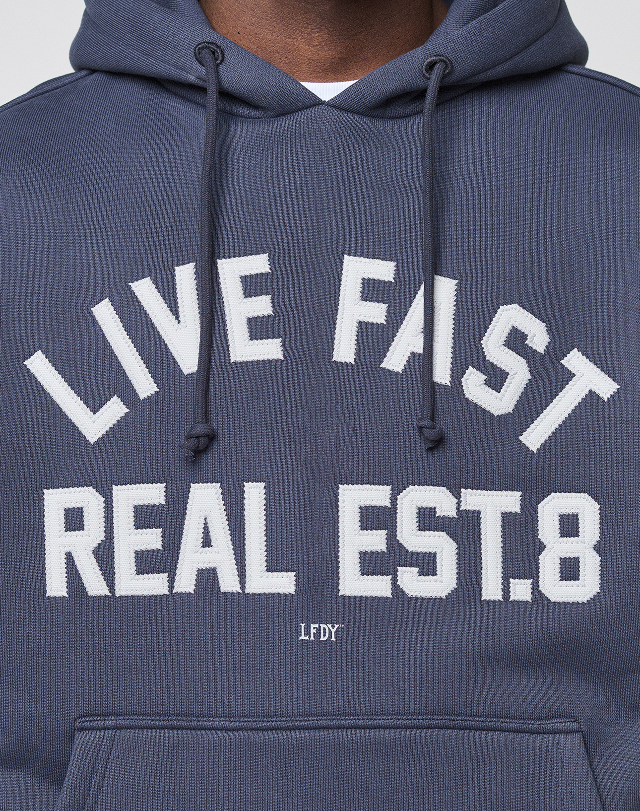A person wears the LIVE FAST DIE YOUNG LFDY Heavyweight Lounge Hooded in dark blue with white "LIVE FAST REAL EST. 8" text, small LFDY lettering below, made from 100% Baumwolle, regular fit; only the upper chest and part of the face are visible.
