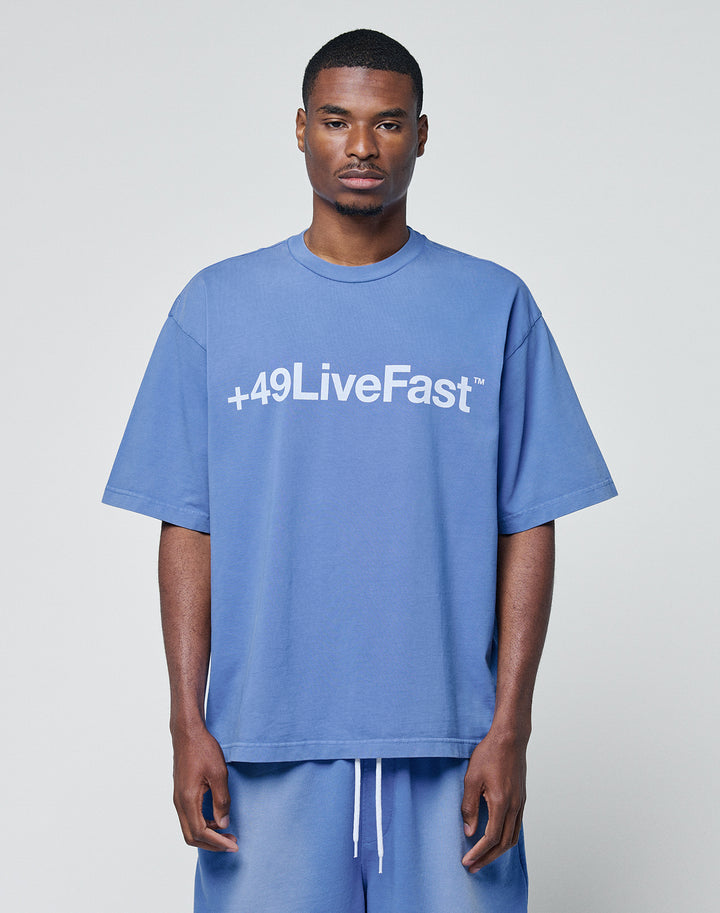 A man faces forward in a blue Basic +49 Tee by LIVE FAST DIE YOUNG, featuring an oversize fit, short sleeves, and bold white +49LiveFast™ print on the front, styled with matching blue shorts against a plain background.