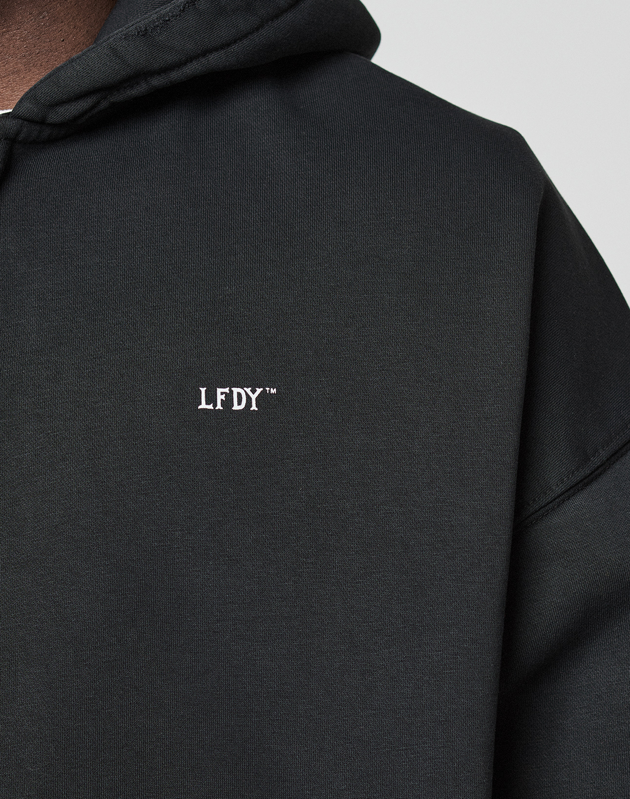 Close-up of the Basic Evidence Hooded Zip by LIVE FAST DIE YOUNG, a black oversized hoodie with bold white branding on the chest. Only part of the wearer’s neck and shoulder shows against a plain light background.