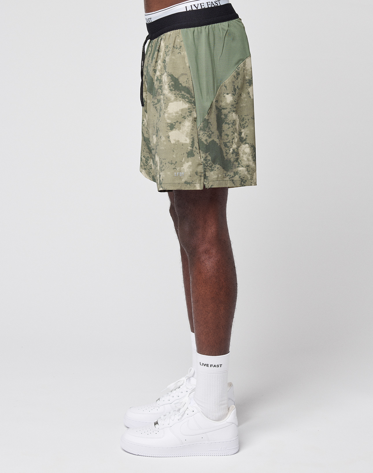 A person stands in profile wearing LIVE FAST DIE YOUNG's LFDY Performance Running Shorts in green camo with a black LIVE FAST waistband, matching white socks, and white Nike sneakers against a plain white background.