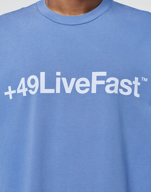 A person wearing an oversize fit blue Basic +49 Tee from LIVE FAST DIE YOUNG featuring +49LiveFast™ printed in white across the chest.