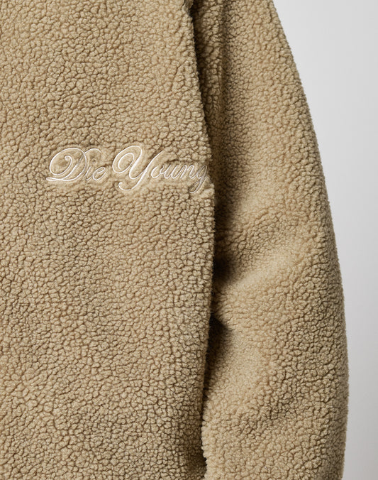 Close-up of the LFDY Teddy Jacket by LIVE FAST DIE YOUNG—a beige, fluffy, oversized jacket with a zipper and “Die Young” embroidered in white cursive on the chest.