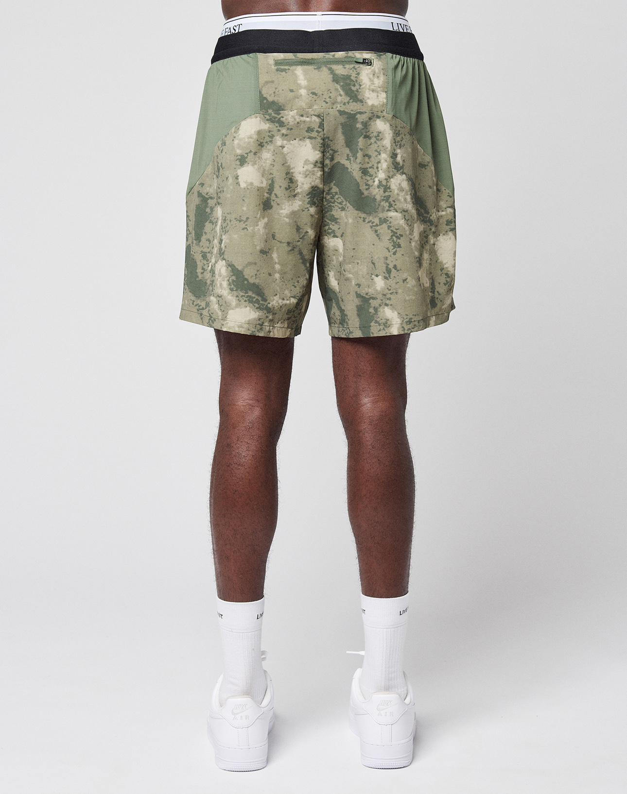 A person stands facing away, wearing green and beige LFDY Performance Running Shorts by LIVE FAST DIE YOUNG with built-in tights, white socks, and white sneakers against a plain light gray background.