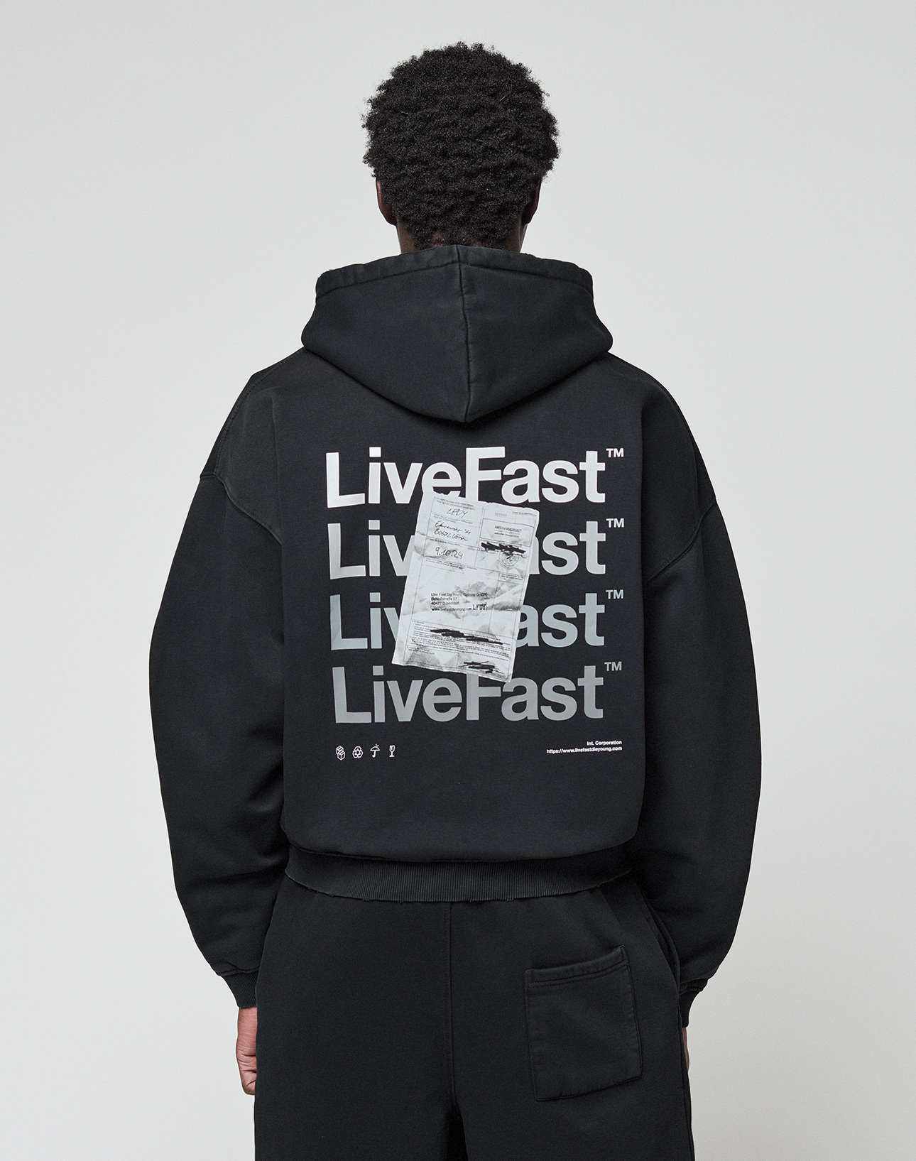 A person wearing the LIVE FAST DIE YOUNG Basic Evidence Hooded Zip in an oversize fit, featuring bold white branding, paper-like graphic, small symbols, and subtle distressing on the back, stands against a plain background.