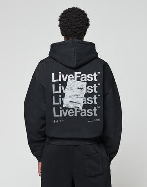 A person wearing the LIVE FAST DIE YOUNG Basic Evidence Hooded Zip in an oversize fit, featuring bold white branding, paper-like graphic, small symbols, and subtle distressing on the back, stands against a plain background.