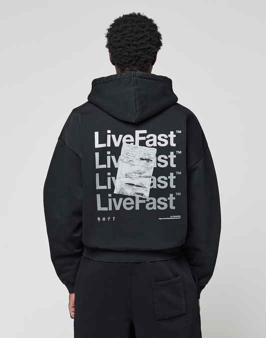 A person wearing the LIVE FAST DIE YOUNG Basic Evidence Hooded Zip in an oversize fit, featuring bold white branding, paper-like graphic, small symbols, and subtle distressing on the back, stands against a plain background.