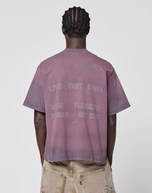 A person with short, twisted hair faces away, wearing the mauve Live Fast District Tee by LIVE FAST DIE YOUNG. This 100% Baumwolle, oversize fit shirt features faded back graphics and text, paired with light pants with visible back pockets. Plain background.