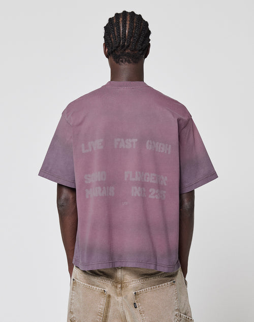 A person with short, twisted hair faces away, wearing the mauve Live Fast District Tee by LIVE FAST DIE YOUNG. This 100% Baumwolle, oversize fit shirt features faded back graphics and text, paired with light pants with visible back pockets. Plain background.