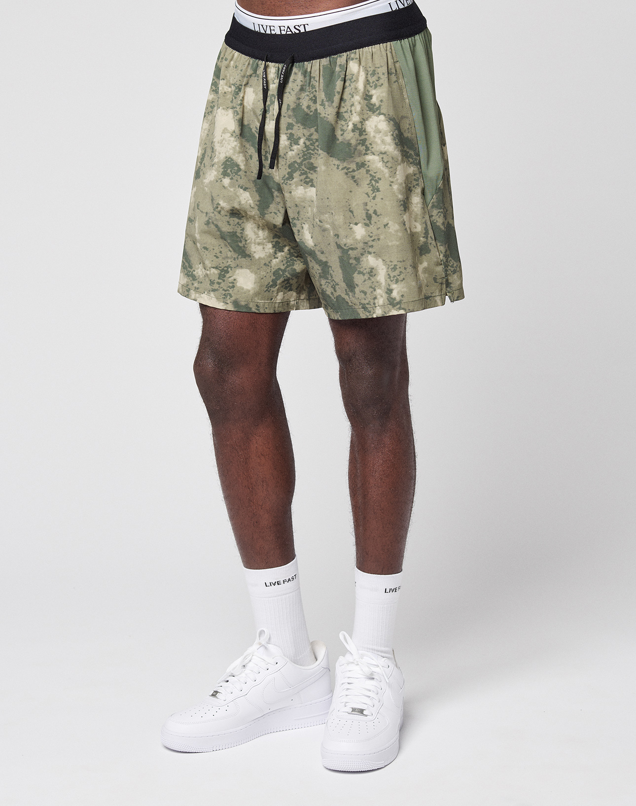 A person wearing LFDY Performance Running Shorts by LIVE FAST DIE YOUNG, featuring green camo print with integrated tights and a white LIVE EAST waistband, paired with white crew socks and sneakers, stands against a plain white background.
