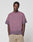 A man with braided hair wears the LIVE FAST DIE YOUNG Live Fast District Tee, an oversized faded purple T-shirt with a subtle spray effect, paired with beige pants, standing hands-in-pockets against a plain white background.