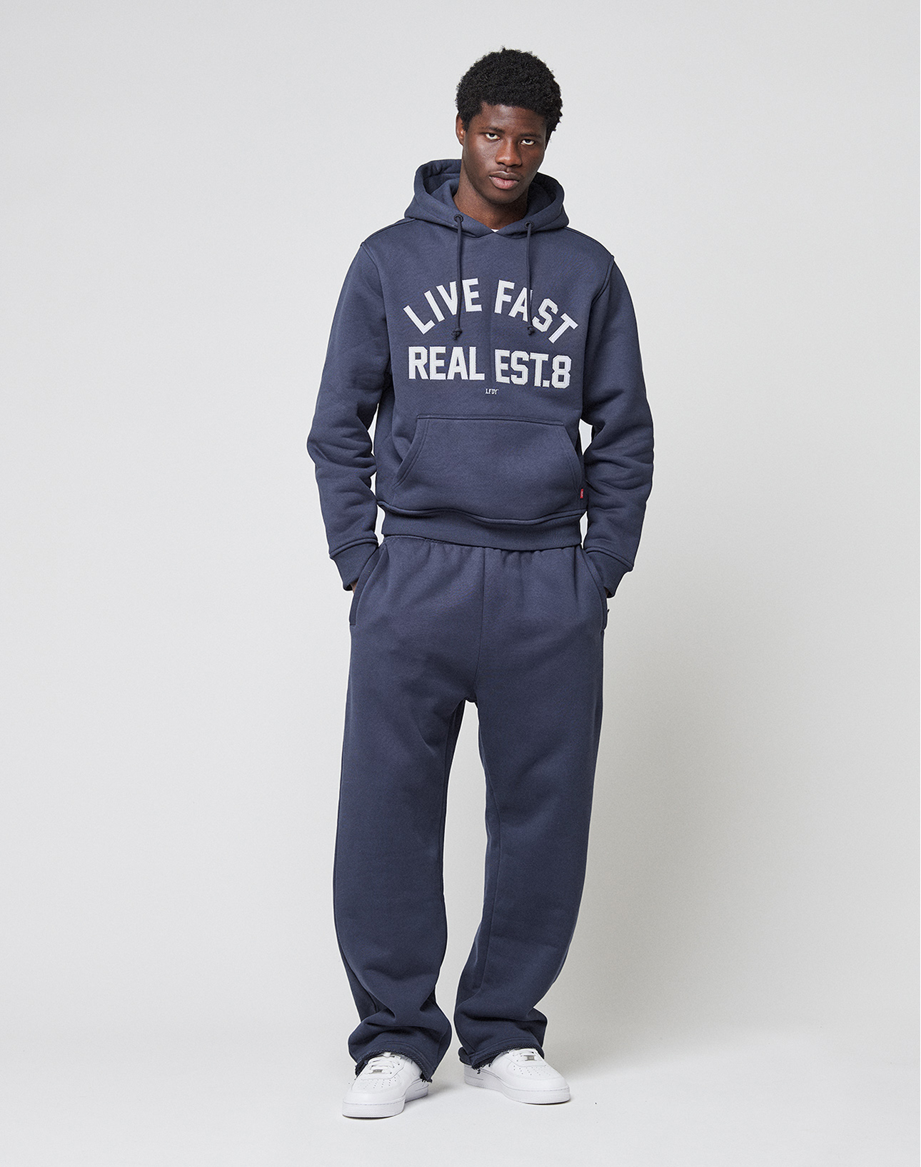 A man stands against a plain background in a navy 650gsm hoodie with LIVE FAST REAL EST. 8, matching LFDY Heavyweight Loungepants by LIVE FAST DIE YOUNG, and white sneakers, with his hands in his pockets.