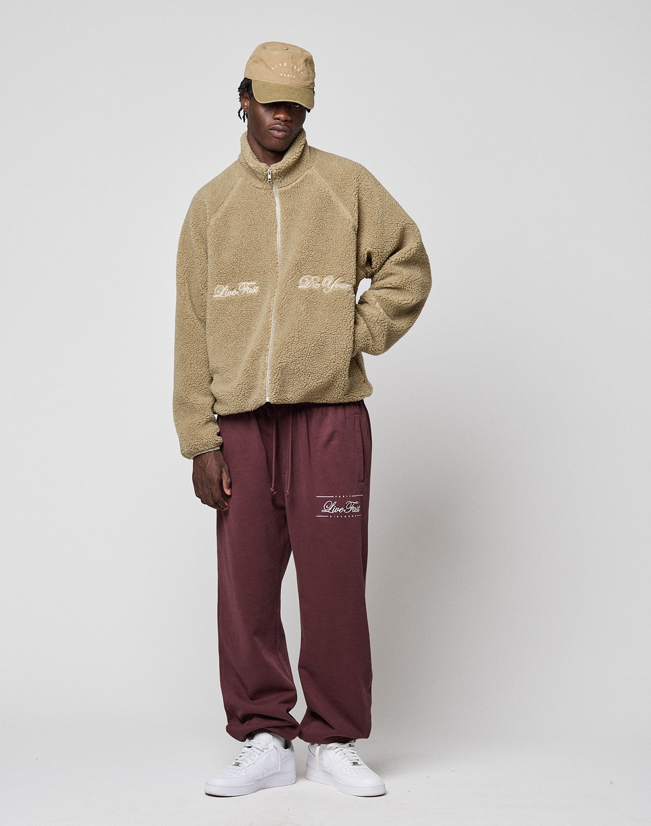 A person in a tan LFDY Teddy Jacket by LIVE FAST DIE YOUNG, burgundy sweatpants, a tan cap, and white sneakers stands relaxed against a plain light background.