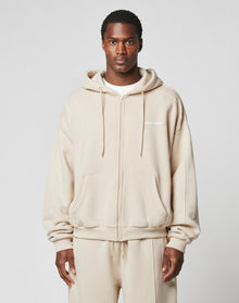 A man faces forward in a LIVE FAST DIE YOUNG LFDY Pleated Hooded Zip, an oversized beige cotton zip-up with hood and front pockets, paired with matching beige pants. The background is plain light gray. | model