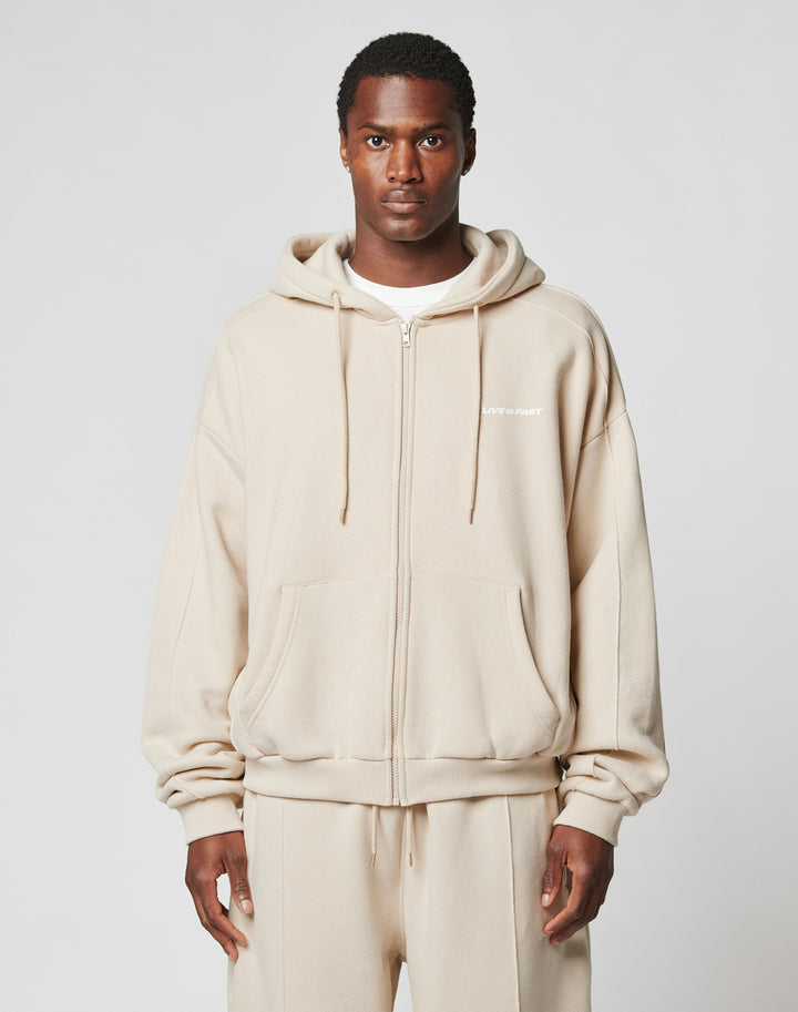 A man faces forward in a LIVE FAST DIE YOUNG LFDY Pleated Hooded Zip, an oversized beige cotton zip-up with hood and front pockets, paired with matching beige pants. The background is plain light gray. | model