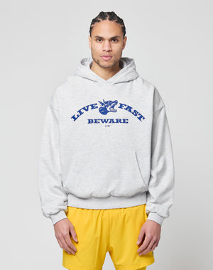 A person with braided hair wears the LIVE FAST DIE YOUNG Live Fast Beware Hooded, a light gray oversized double-layer hoodie with a bold front print, styled with blue and yellow shorts against a plain light background.