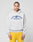A person with braided hair wears the LIVE FAST DIE YOUNG Live Fast Beware Hooded, a light gray oversized double-layer hoodie with a bold front print, styled with blue and yellow shorts against a plain light background.
