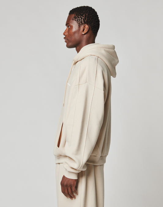 A man in profile wears the LIVE FAST DIE YOUNG LFDY Pleated Hooded Zip in an oversized beige fit with matching sweatpants against a plain, light background.