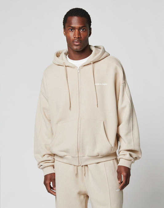 A man wearing an LFDY Pleated Hooded Zip by LIVE FAST DIE YOUNG and matching beige sweatpants, both made from cotton, stands facing forward against a plain light gray background.