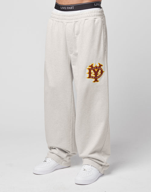 A person stands against a plain white background wearing LIVE FAST DIE YOUNG DY Jogger sweatpants in light beige with a yellow and red emblem on the left thigh, white sneakers, and black “LIVE FAST” logo underwear.