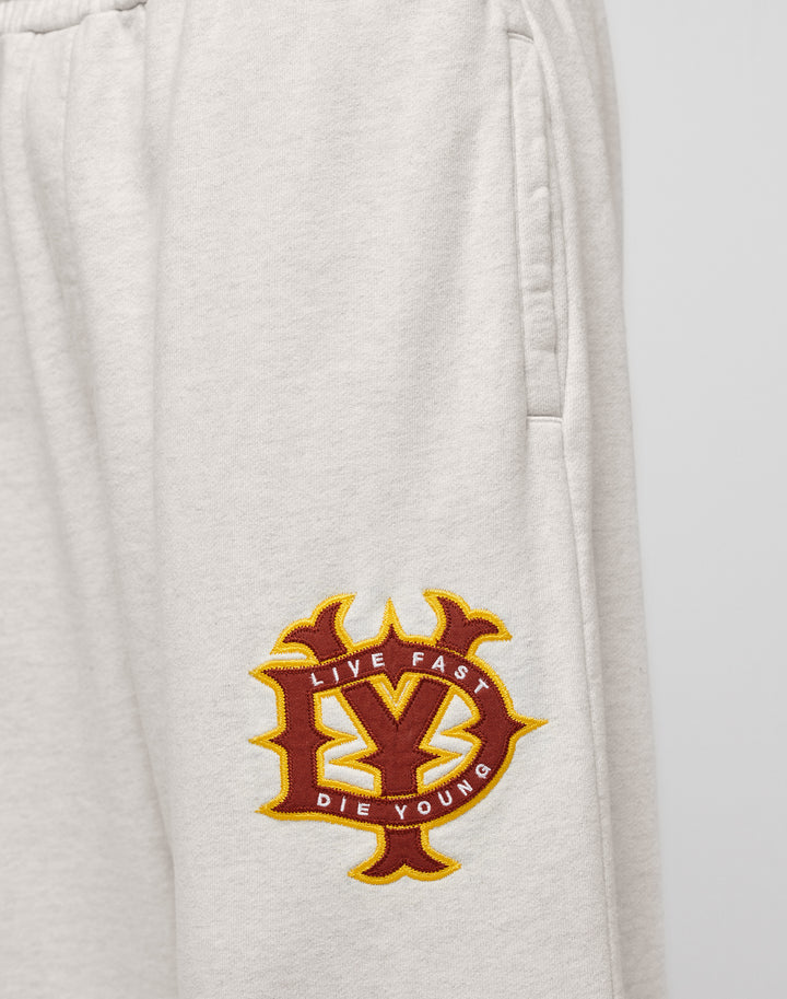 Close-up of DY Jogger by LIVE FAST DIE YOUNG: beige, oversized sweatpants with a maroon and yellow LY patch near the pocket reading “LIVE FAST DIE YOUNG.” Made from 100% cotton for all-day comfort.