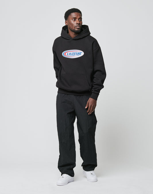 A man stands against a plain white background wearing the oversized fit Live Fast Garment Hooded by LIVE FAST DIE YOUNG, paired with black pants and white sneakers. He looks slightly left with his hands by his sides.