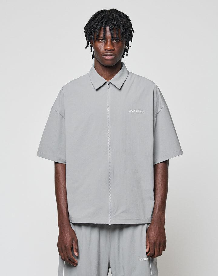 A man with short dreadlocks wears the LIVE FAST DIE YOUNG LFDY Ripstop Shirt and matching gray shorts, both with bold LIVE FAST Silikonprint text, standing against a plain light background. | model