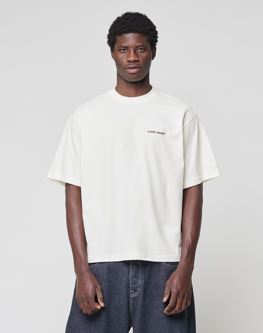 A man stands facing forward against a plain background, wearing the LIVE FAST DIE YOUNG LFDY Grand Theft Tee in white with front and back print, paired with dark blue jeans. The tee is made from 100% Baumwolle; his arms are relaxed at his sides.