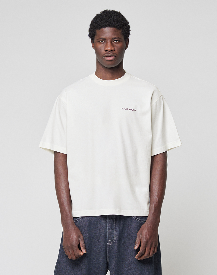 A man stands facing forward against a plain background, wearing the LIVE FAST DIE YOUNG LFDY Grand Theft Tee in white with front and back print, paired with dark blue jeans. The tee is made from 100% Baumwolle; his arms are relaxed at his sides.
