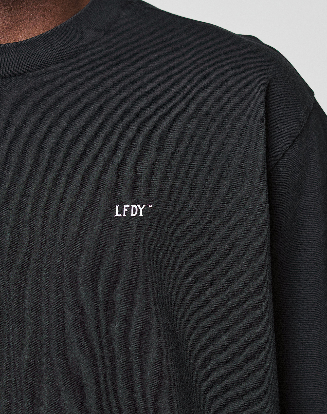 A close-up of a person wearing the LIVE FAST DIE YOUNG Basic Evidence Tee, featuring a plain black oversized fit and small white “LFDY™” text embroidered on the chest, focusing on the upper torso and shoulder.