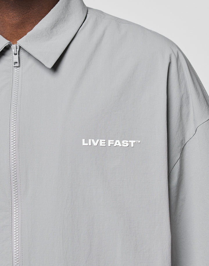 Close-up of a person wearing the LIVE FAST DIE YOUNG LFDY Ripstop Shirt in light gray with an oversize fit and collar. LIVE FAST™ is printed in white silicone on the left chest. Only part of their neck and shoulder are visible.