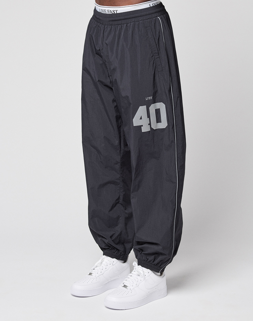 A person wears LIVE FAST DIE YOUNG LFDY Performance Trackpants in black, made of 100% nylon with a grey LIVE 40 reflektierender print on the left thigh, paired with white sneakers. Only the lower body is shown against a white background.