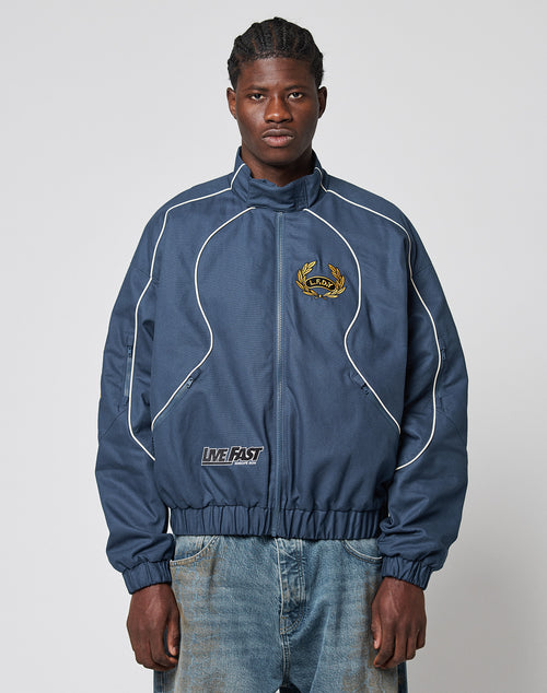 A man wearing an LFDY Canvas Jacket by LIVE FAST DIE YOUNG with white piping, multi-logo embroidery, elastic cuffs, and baggy blue jeans stands against a simple light background, looking forward.