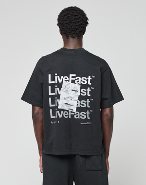 A person, facing away from the camera, wears the LIVE FAST DIE YOUNG Basic Evidence Tee—an oversize black 100% Baumwolle shirt with a bold “LiveFast” backprint in white and an overlapping paper graphic.