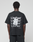 A person, facing away from the camera, wears the LIVE FAST DIE YOUNG Basic Evidence Tee—an oversize black 100% Baumwolle shirt with a bold “LiveFast” backprint in white and an overlapping paper graphic.