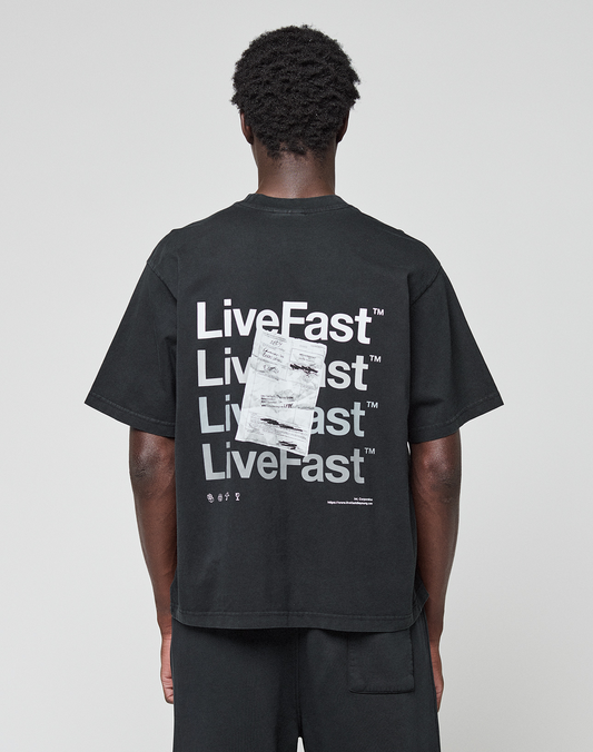 A person, facing away from the camera, wears the LIVE FAST DIE YOUNG Basic Evidence Tee—an oversize black 100% Baumwolle shirt with a bold “LiveFast” backprint in white and an overlapping paper graphic.