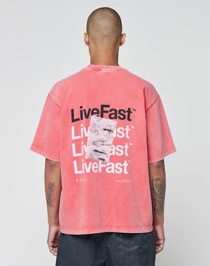 A person with a shaved head faces away, wearing the LIVE FAST DIE YOUNG Basic Evidence Tee in faded red. The back features repeated bold white and black LiveFast prints, a graphic, small white text below, and a subtle flag label on the hem.