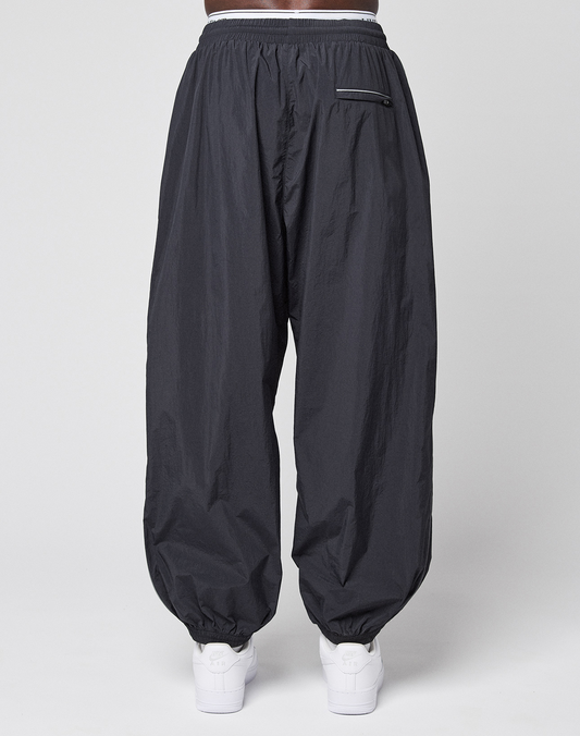 A person wears LIVE FAST DIE YOUNG's LFDY Performance Trackpants in black, featuring an oversize fit, 100% nylon material, elastic waistband and cuffs, and a zippered back pocket. White sneakers complete the look against a plain white background.