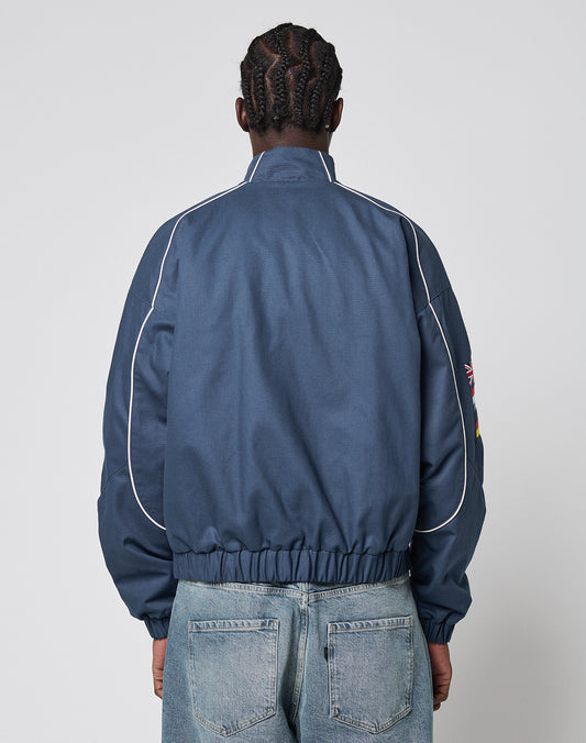 A person with braided hair is seen from behind, wearing the LFDY Canvas Jacket by LIVE FAST DIE YOUNG—blue with multilogo embroidery, white piping, elastic cuffs, and a high collar—paired with light blue jeans against a plain light background.