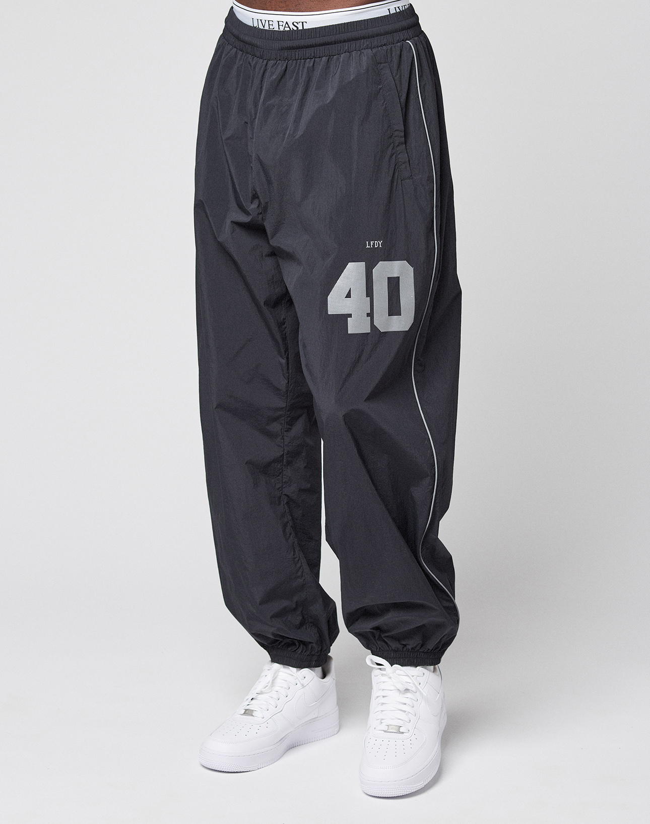 Someone wearing the oversize fit LFDY Performance Trackpants by LIVE FAST DIE YOUNG in black with a white "40" and "LIVE FAST" on the waistband and thigh, plus a reflective print, styled with white sneakers against a plain light background.