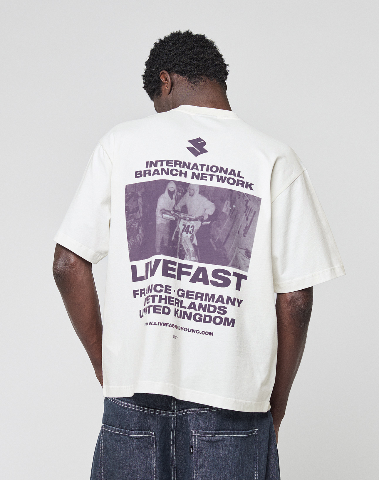 A person wearing the LFDY Grand Theft Tee by LIVE FAST DIE YOUNG—an oversize white 100% cotton shirt with bold front and back prints. The back displays "INTERNATIONAL BRANCH NETWORK," "LIVEFAST," several countries, and a vintage race photo in purple.