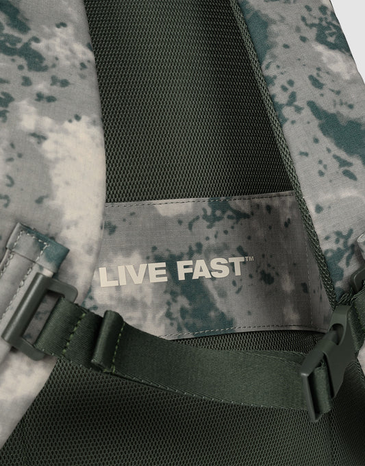 Close-up of the LFDY Travel Backpack by LIVE FAST DIE YOUNG, a green and gray camo 25L rucksack featuring a mesh panel, adjustable shoulder straps, and an inner label reading "LIVE FAST" in bold white letters.