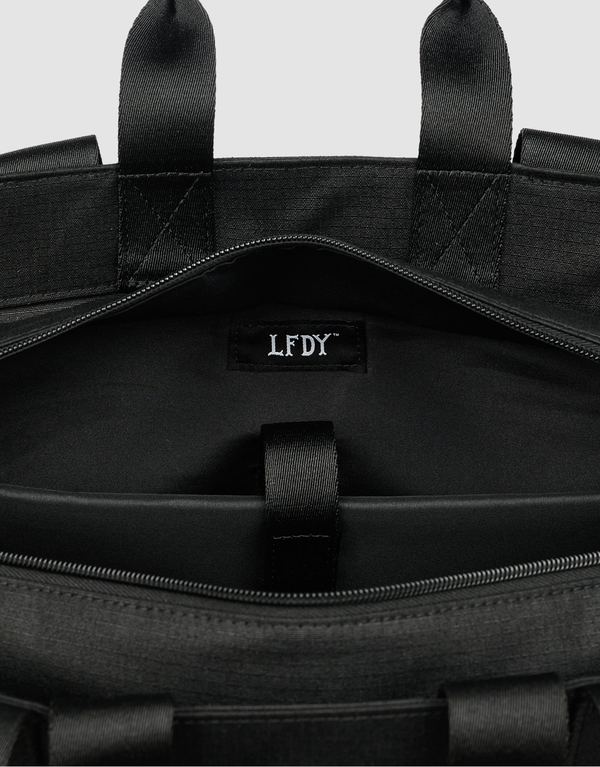 Close-up of the inside of a black LFDY Utility Bag by LIVE FAST DIE YOUNG, highlighting a fabric tag with white LFDY lettering. The bag includes black straps, a looped fastener near the opening, and a large compartment for extra storage.